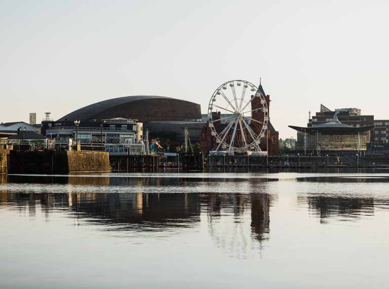 Golygfa o Fae Caerdydd o’r dŵr sy’n dangos adeiladau ac olwyn fawr