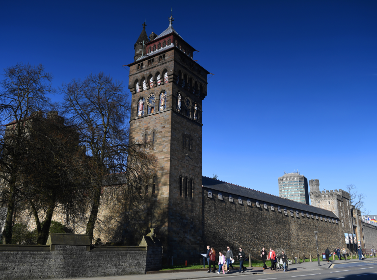 Muriau cerrig a thŵr cloc Castell Caerdydd, gyda phobl yn cerdded ar y palmant o dan awyr las glir.
