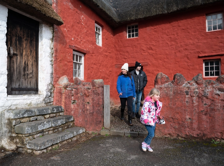 Oedolyn a dau o blant tu allan i ffermdy wedi ei baentio’n goch.