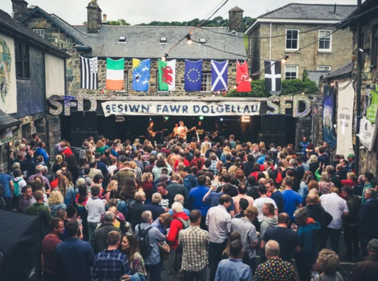 Cynulleidfa fawr yn gwylio band gwerin Calan ar lwyfan Sesiwn Fawr Dolgellau. Mae sawl fflag uwch ben baner Sesiwn Fawr gan gynnwys Cymru, Ewrop, Yr Alban ac Ynys Manaw.