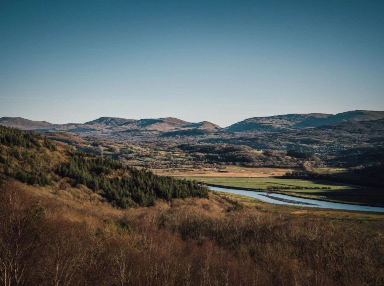 Golygfa o fynyddoedd a choetir gwyrdd gydag aber yr afon Mawddach yn y canol. Mae'r awyr yn las ar ddiwrnod braf.