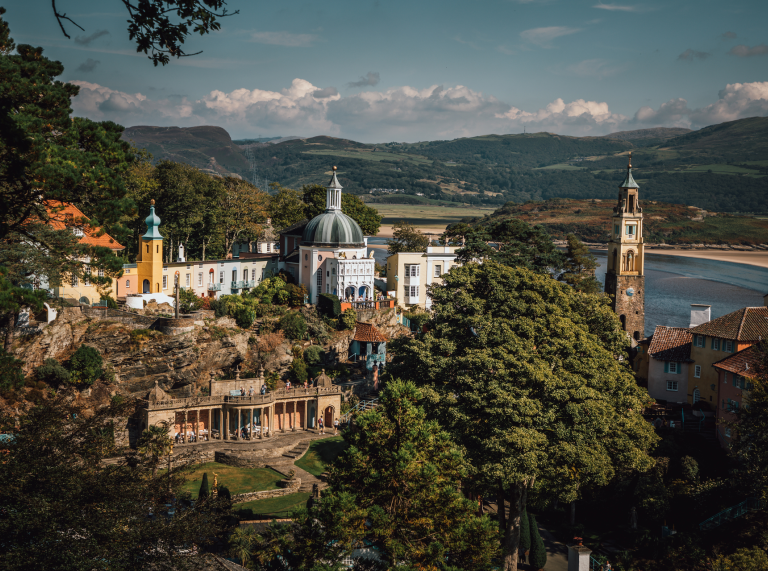Golygfa o bentref Portmeirion o’r awyr gyda’r môr a’r mynyddoedd yn y cefndir.
