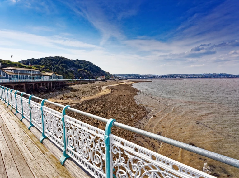 rhan o'r llwybr estyllod a chanllaw y pier, gyda golygfa o draeth a bryn yn y cefndir.