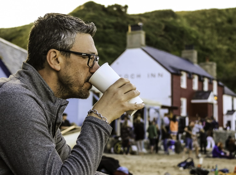 Dyn yn cael diod y tu allan i dafarn ar y traeth.