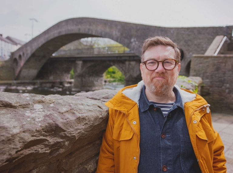 Dyn (Gareth Potter) mewn cot felen o flaen pont fwa cerrig.