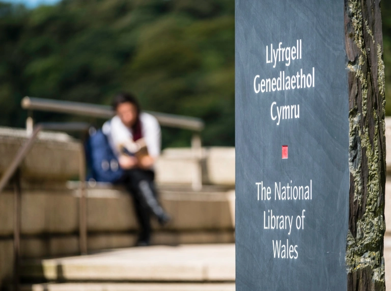 Arwydd lechen Llyfrgell Genedlaethol Cymru gyda pherson yn eistedd yn y cefndir