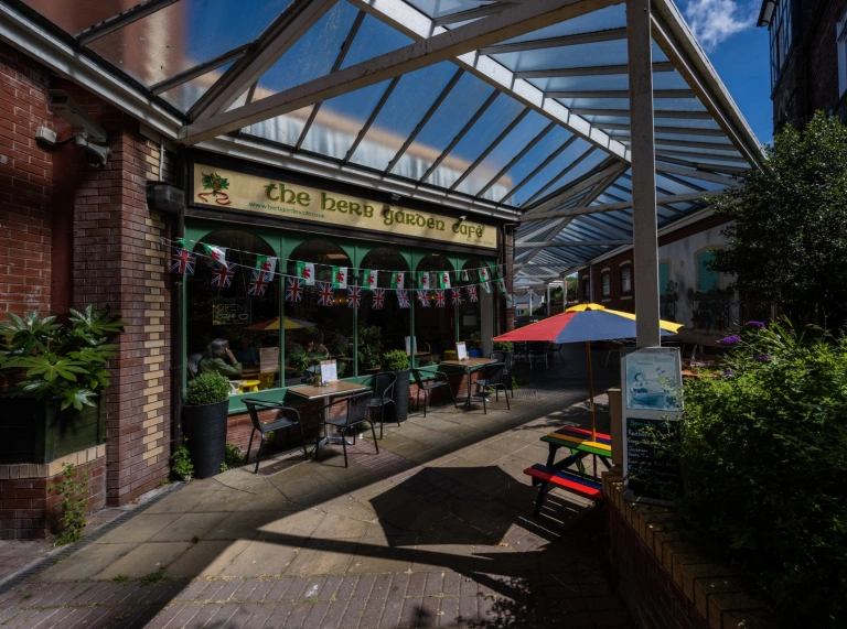 Blaen caffi dan lwybr cerdded wedi’i orchuddio â gwydr, gyda seddi lliwgar y tu allan.