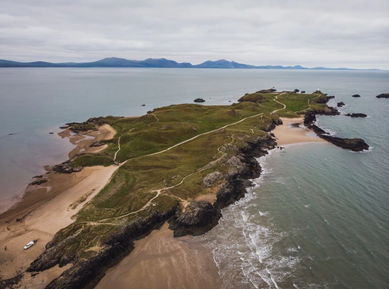 Penrhyn glaswelltog gyda thraethau euraidd a goleudy gwyn.