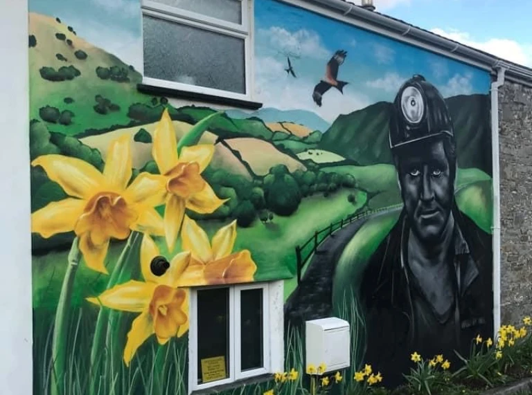 Golygfa allanol o furlun ar gefn y bwthyn yn darlunio glöwr, cenhinen Bedr a bryniau gwyrdd tonnog.