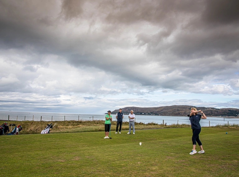 Grŵp o bobl yn chwarae golff ar gwrs golff arfordirol.