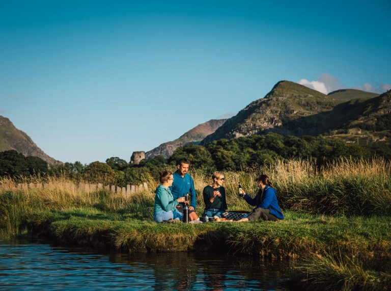 Teulu’n cael picnic wrth ochr llyn.