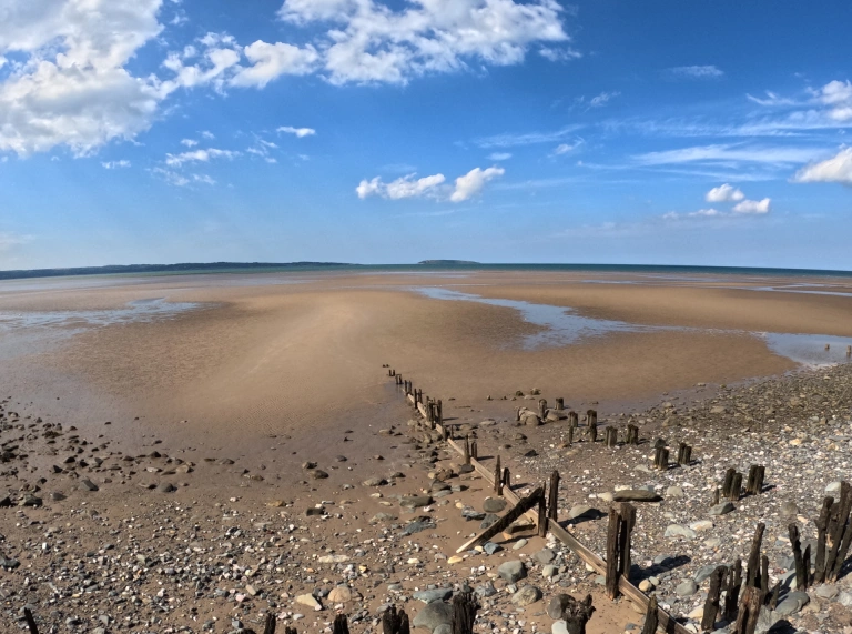 Golygfa ar draws traeth tywodlyd allan i'r môr gyda'r llanw allan ac awyr las uwchben.