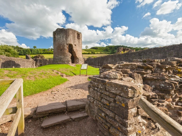 Adfeilion cerrig castell canoloesol, gyda thŵr crwn a muriau isel gerllaw llwybr glaswelltog o dan awyr rannol gymylog.