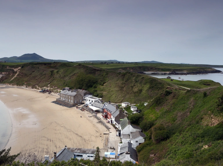 Llun o adeiladau ar y traeth, gyda bryniau gwyrdd y tu ôl.
