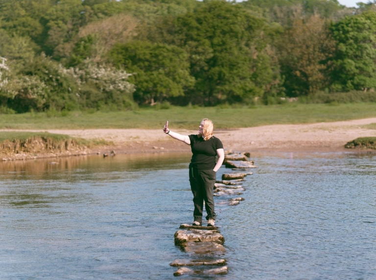 Person mewn Person yn sefyll ar gerrig mewn afon yn tynnu hunlun.