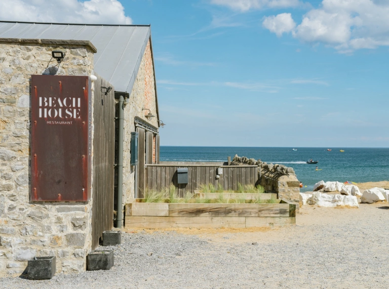 Tu allan bwyty Beach House yng Ngŵyr gyda'r môr yn y cefndir.
