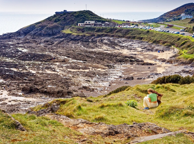 Bae creigiog ger y môr, gyda chlogwyni isel a llefydd parcio bob ochr iddo.