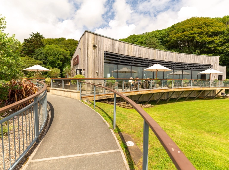 Llwybr a rheiliau sy'n arwain at adeilad modern gyda bwyty a seddi allanol.