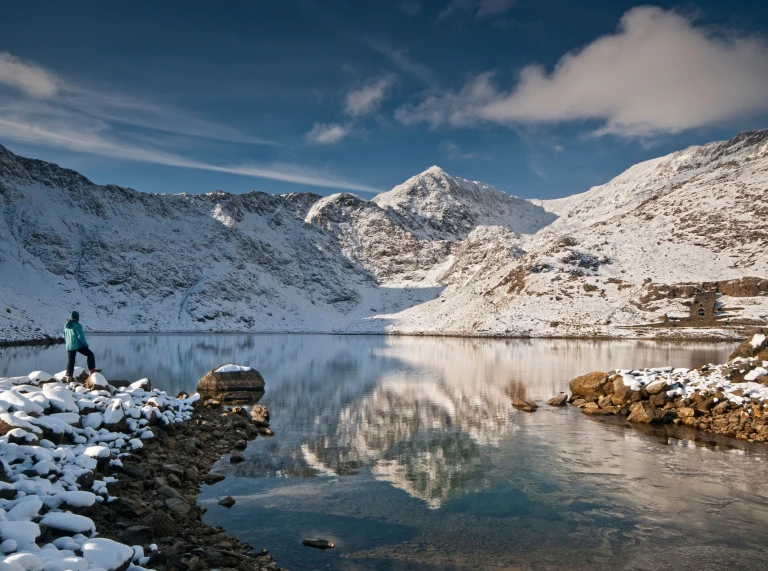 Cerddwr yng nghanol copaon o eira yn Eryri - ar lan Llyn Llydaw yn y gaeaf.