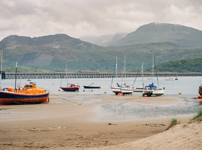 Cychod ar draeth gyda môr a mynyddoedd yn gefndir.