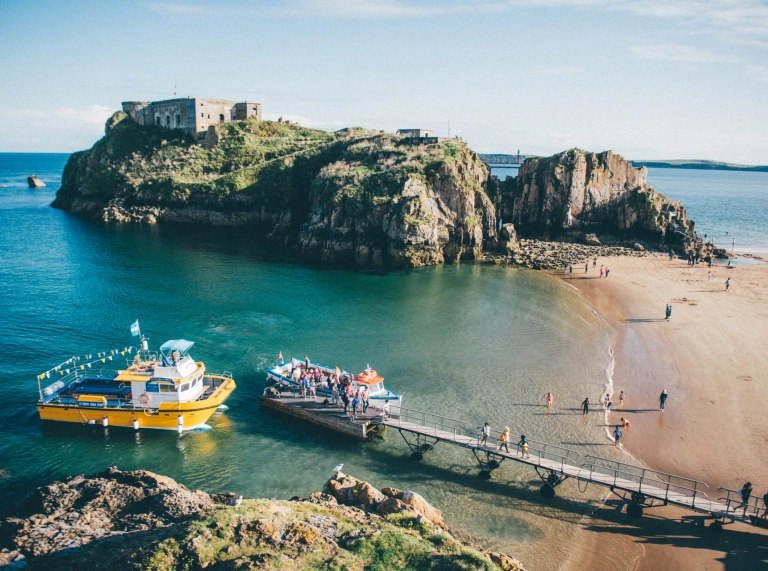 Llun o Gaer Ynys Catrin ar Ynys Catrin, a chychod bach yn casglu teithwyr o Ddinbych-y-pysgod