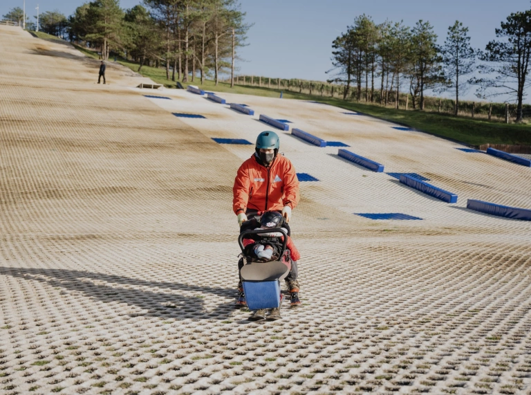 Plentyn mewn sled / pram sgïo wedi’i addasu yn cael ei wthio gan ddyn ar lethr sgïo sych.