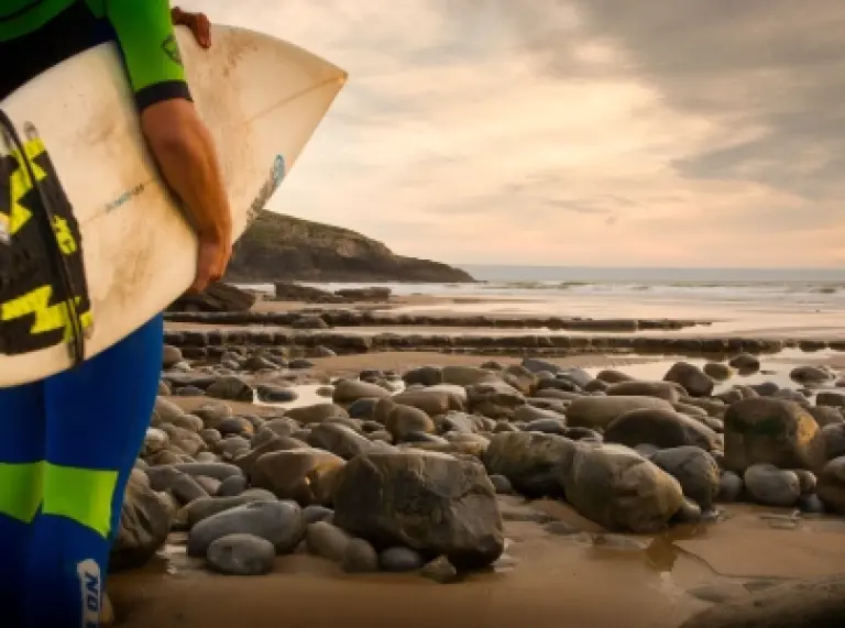 Llun o berson gyda'u cefn at y camera, yn wynebu'r traeth a'r môr, gyda bwrdd syrffio dan eu cesail