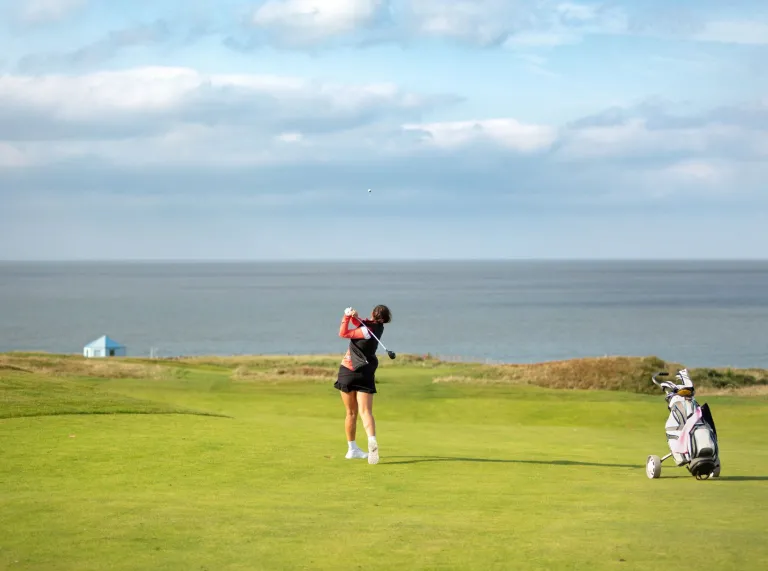 Golffiwr yn sefyll ar y gwyrdd, yn cymryd siglen. Gellir gweld y môr y tu hwnt i wyrdd y cwrs golff.