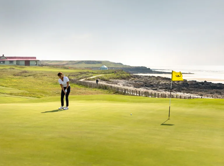 Golffwr yn sefyll ar y green, yn rhoi i'r twll gyda baner ynddo. Mae adeilad y clwb yn weladwy yn y pellter o atrás.
