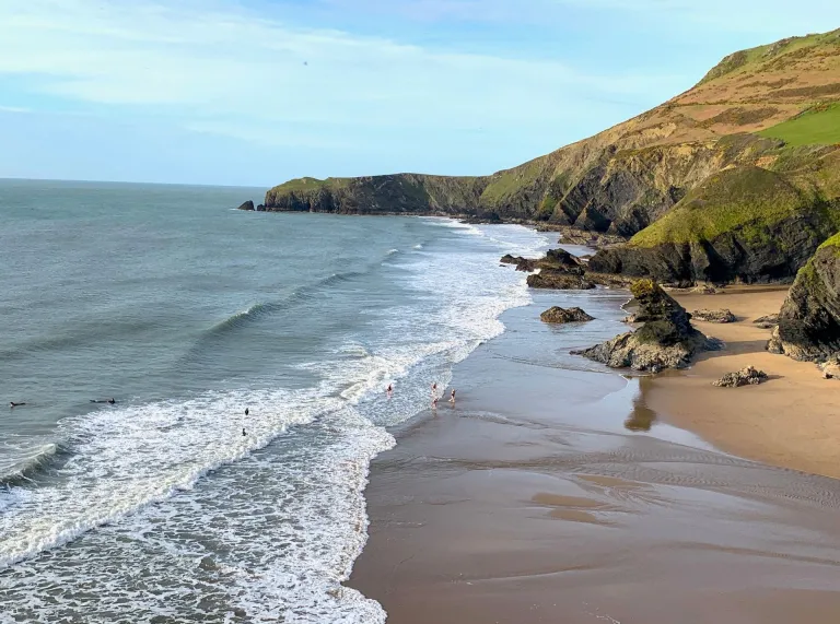 Traeth tywodlyd gyda thonnau, clogwyni creigiog yn y cefndir, ac ychydig o bobl yn mwynhau’r dŵr o dan awyr fymryn yn gymylog.