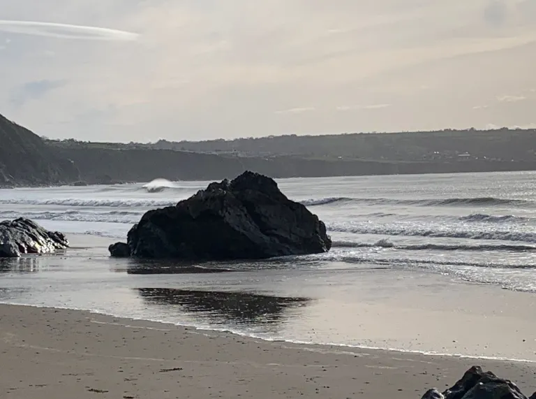 Craig wedi’i gorchuddio’n rhannol gan y lli ar draeth tawel gyda chlogwyni yn y pellter ac awyr gymylog.