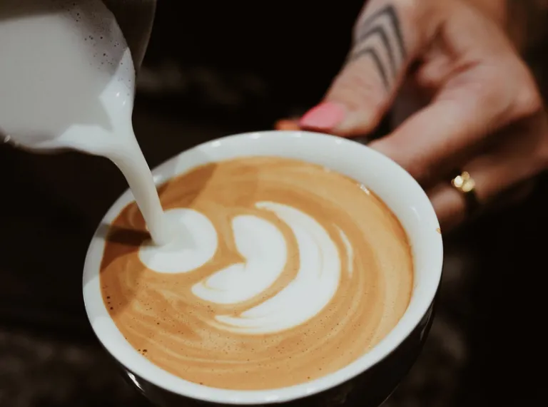 Close-up o laeth stemio yn cael ei dywallt i mewn i baned o goffi i greu celf latte a llaw gyda tatŵ a modrwy.