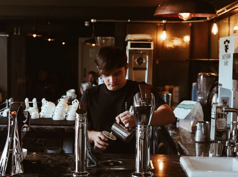 Mae barista yn paratoi coffi y tu ôl i gownter caffi tywyll, diwydiannol gyda tapiau crôm a goleuadau crog cynnes.