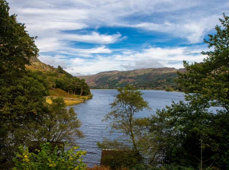 Edrych dros lyn sy’n llechu rhwng bryniau coediog.