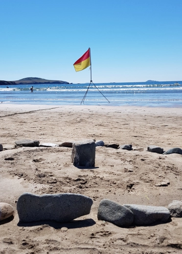 creigiau mewn cylch ar dywod, gyda baner a môr yn y cefndir.