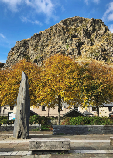 Carreg fawr yng nghanol tref Blaenau Ffestiniog, gyda chraig fawr yn y cefndir ac awyr las.