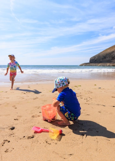 Plentyn yn adeiladu castell dywod a phlentyn yn y cefndir yn chwarae.
