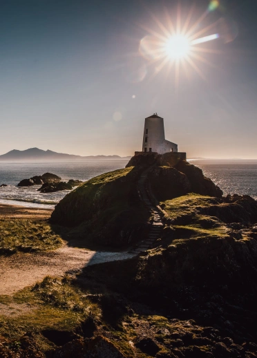 Goleudy gwyn ar ynys. Mae'r haul yn disgleirio yn y cefndir.