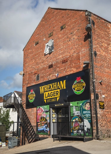 Adeilad brics coch gydag arwydd Wrexham Lager mewn du uwchben y drws.