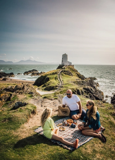 Teulu’n cael picnic gyda goleudy yn y cefndir.