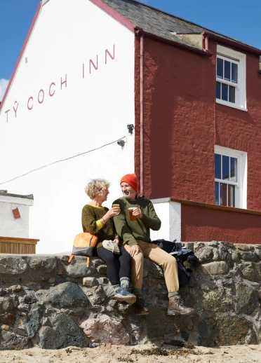 Cwpwl yn eistedd ar wal yn mwynhau diod y tu allan i Dafarn Ty Coch