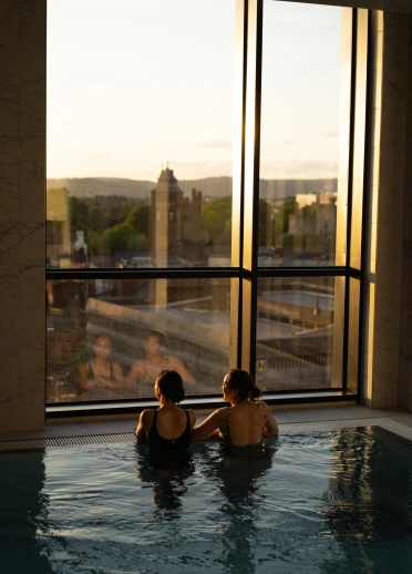 Two people in an infinity pool looking out of the window at a cityscape view with setting sun.