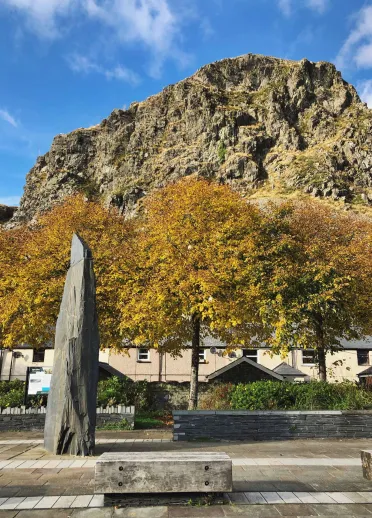Carreg fawr yng nghanol tref Blaenau Ffestiniog, gyda chraig fawr yn y cefndir ac awyr las.