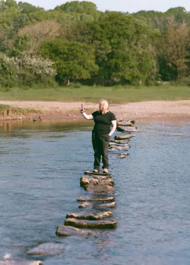 Dŵr gyda cherrig sarn yn ei groesi. Oedolyn yn tynnu hunlun wrth sefyll ar y cerrig. Llwyni yn y cefndir.