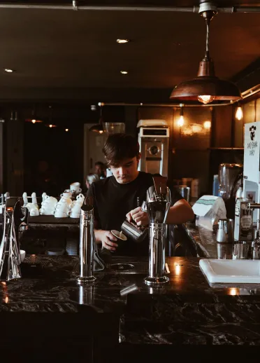 Mae barista yn paratoi coffi y tu ôl i gownter caffi tywyll, diwydiannol gyda tapiau crôm a goleuadau crog cynnes.
