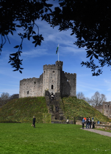 People walking up to The Keep, a tower at Cardiff Castle framed by a tree in the foreground.