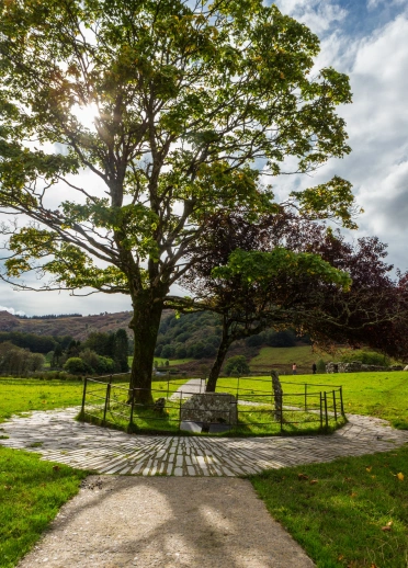 Llwybr gro mân yn arwain at goeden wrth garreg goffa