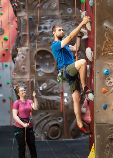 Dyn yn dringo ar wal ddringo dan do gyda menyw’n dal y rhaff.