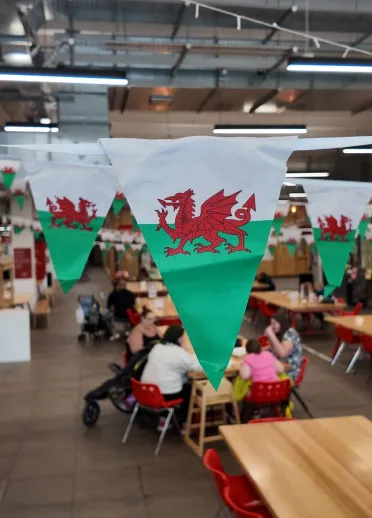 Ardal fwyta fywiog gyda baneri bach Baner Cymru yn addurno'r gofod, a phobl yn ymlacio wrth fyrddau a chadeiriau.