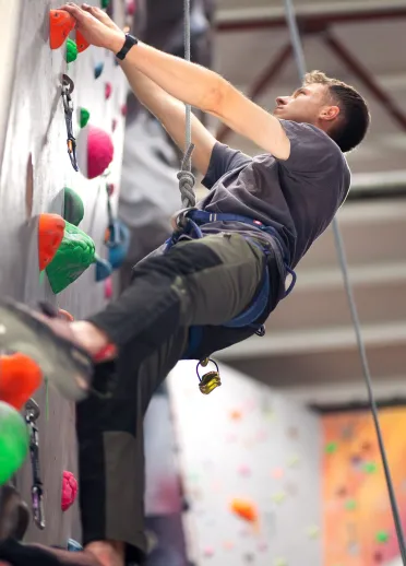 Dringwr yn dringo wal dan do gyda gafaelion gwyrdd, oren a phinc, ac yn gwisgo offer diogelwch a harnais.